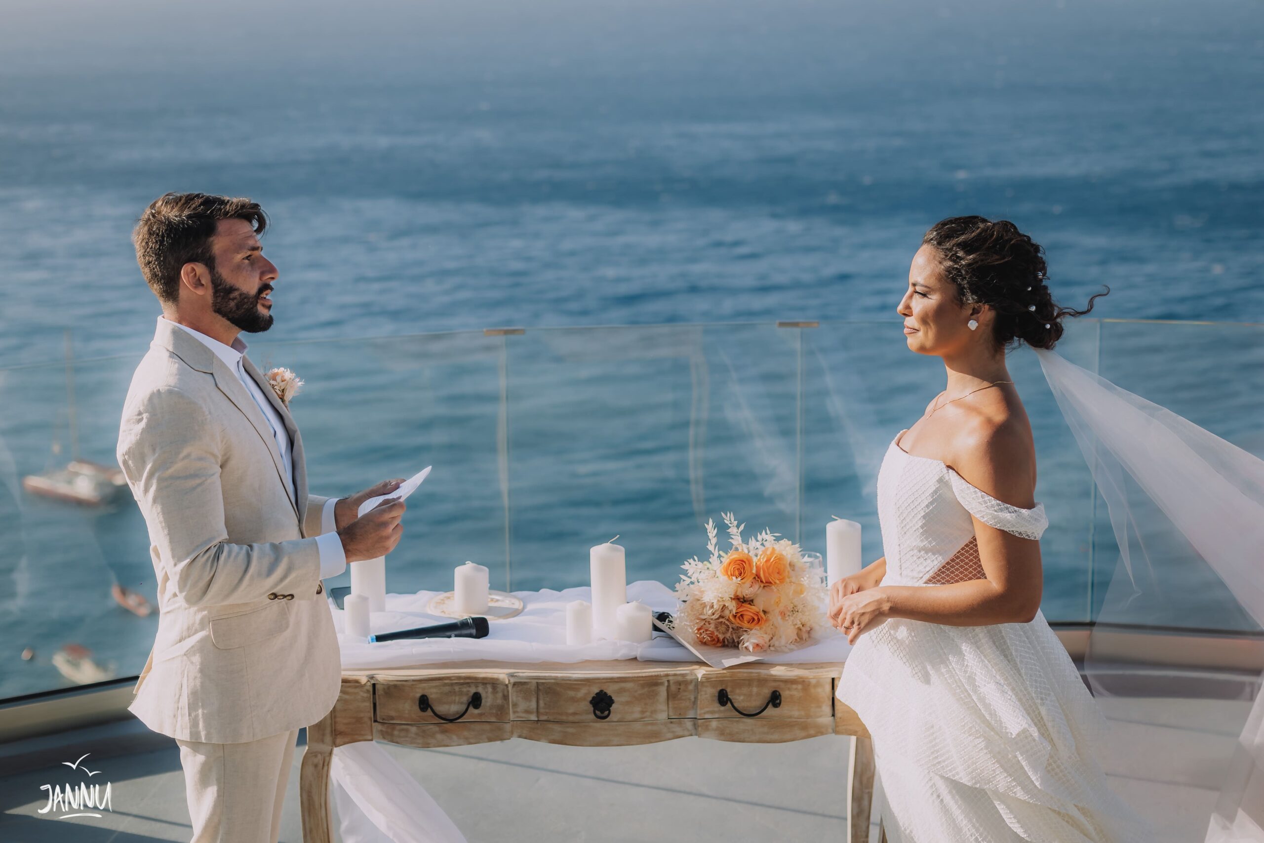 Casal trocando votos em casamento em Santorini, Grécia. Noivo vestido com terno bege e noiva vestida com vestido branco.
