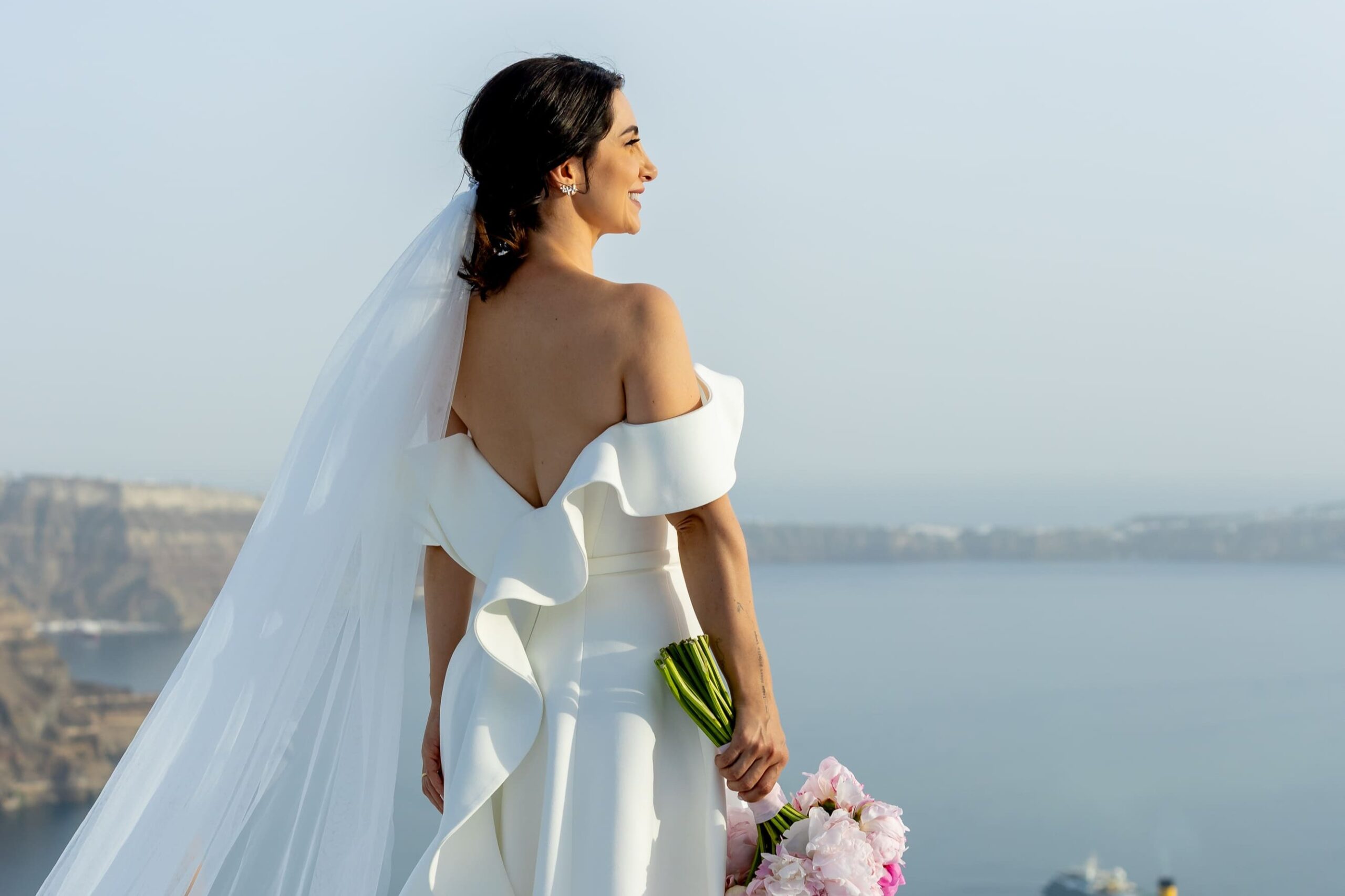 Noiva vestida de branco, segurando um buque de rosas, observando a paisagem da caldeira em Santorini, Grécia