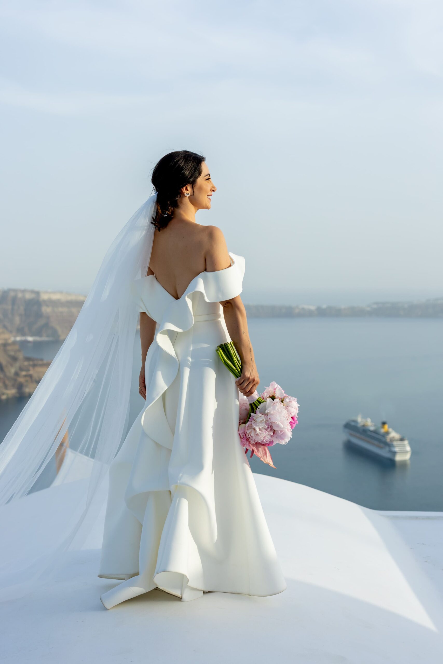Noiva vestida de branco, segurando um buque de rosas, observando a paisagem da caldeira em Santorini, Grécia