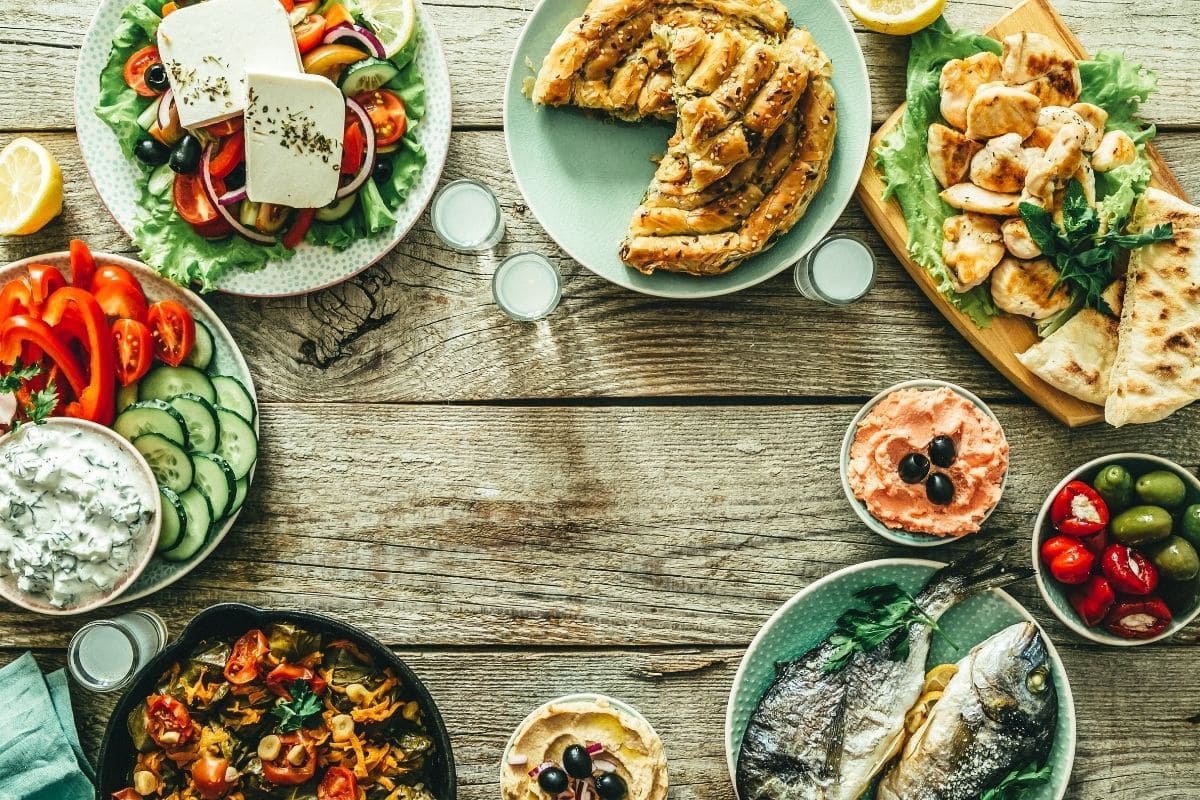 Os sabores da culinária grega com pratos típicos como salada grega, peixe fresco, frango grelhado, torta tradicional, azeite, ervas mediterrâneas e mezes autênticos.