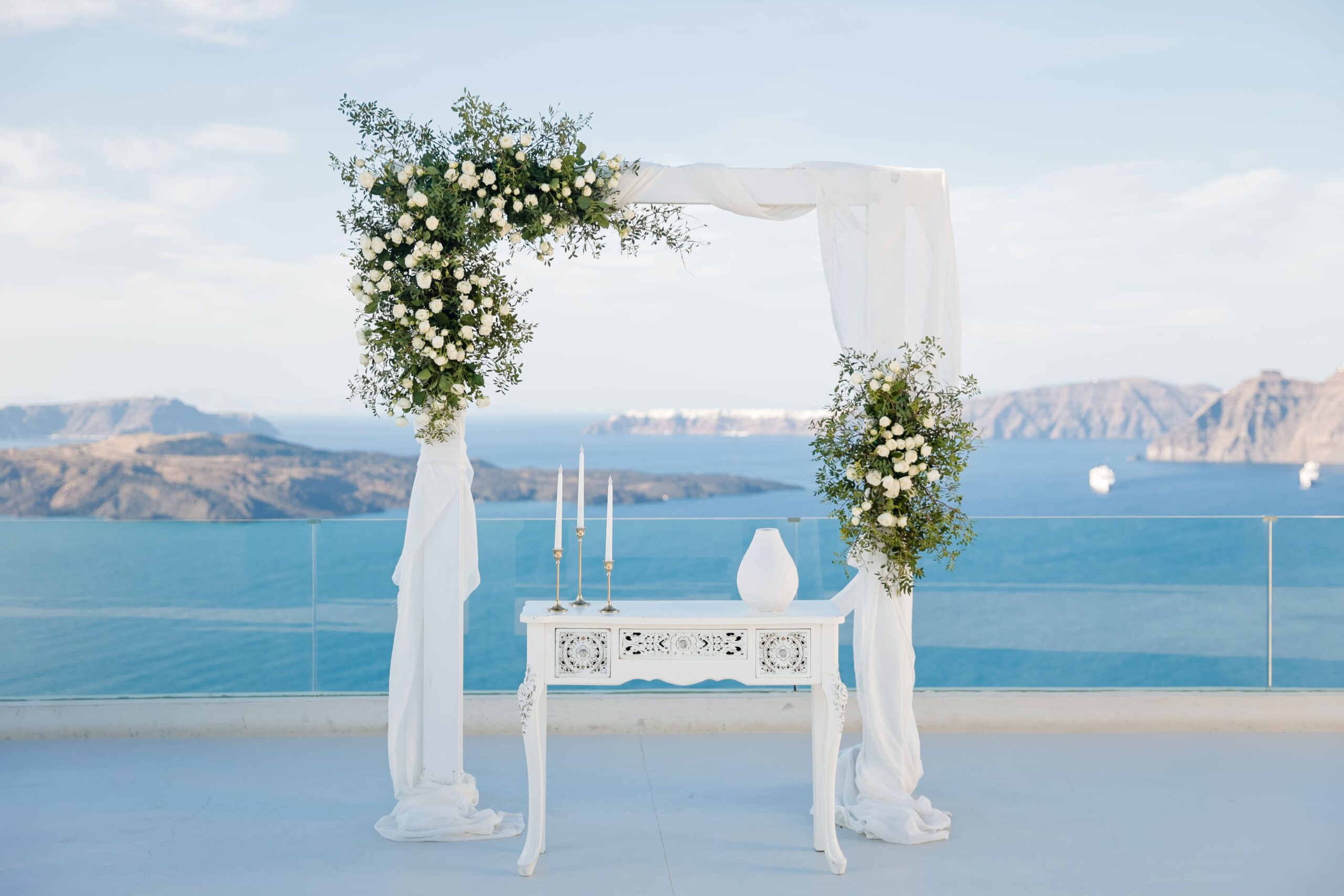 Decoração gazebo com dois arranjos, mesa ao centro, com velas e vaso, vista da caldeira ao fundo, em Santorini, Grecia