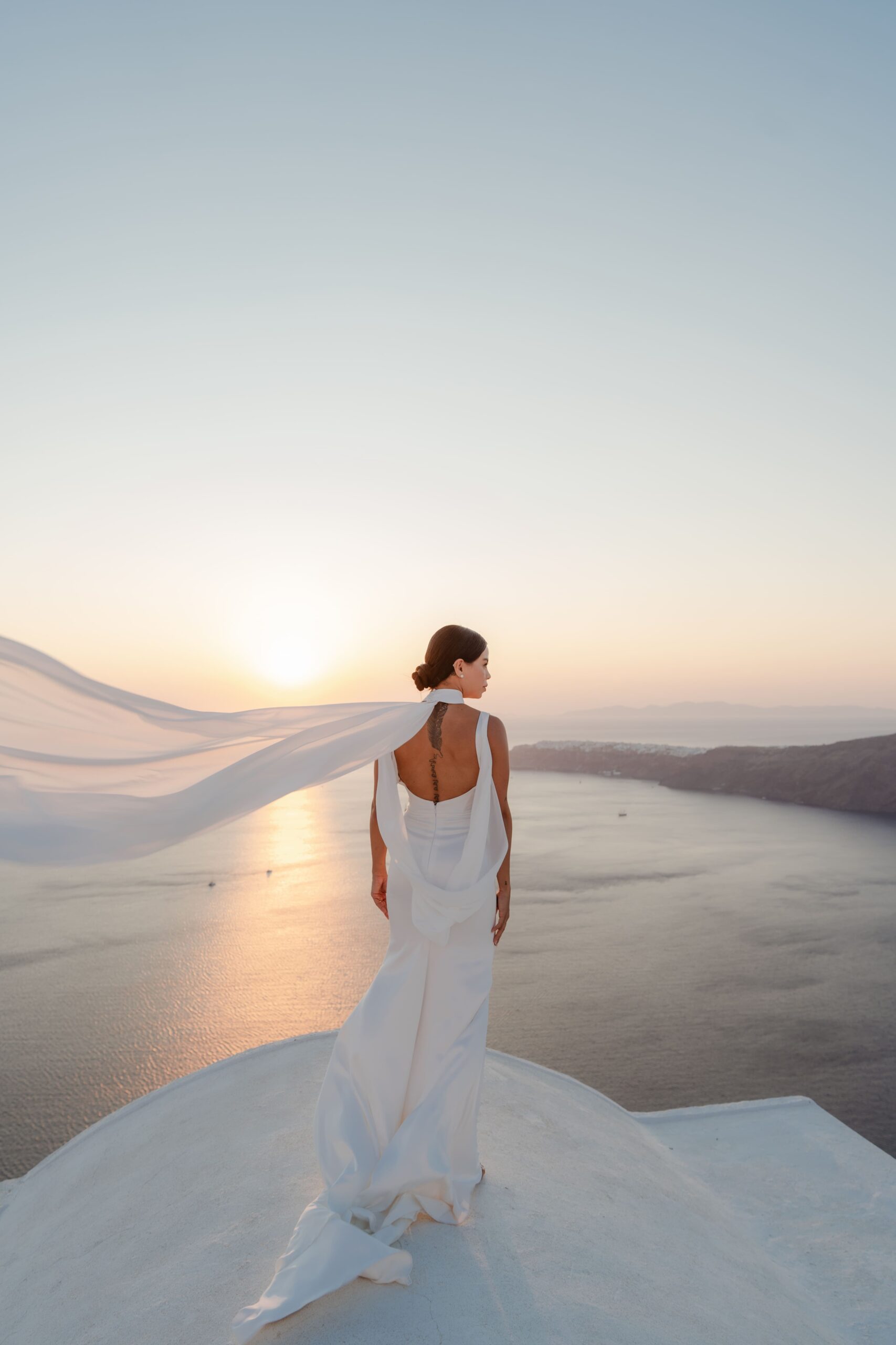 Noiva, com vestido branco, de costas, observando a caldeira, em um por do sol em Santorini, Grecia